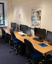 3 Arbeitsplätze mit jeweils 2 Bildschirmen, einem Bürostuhl, Tastatur, Maus und Headset. An der Wand ein Kalender und ein WBS TRAINING Plakat.