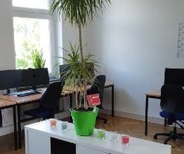 Schulungsraum mit mehreren Arbeitsbereichen, mit 2 PC-Bildschirmen, Tastatur, Maus und Headset ausgestattet sind. Im Vordergrund steht ein weißes Regal mit schwarzen Fächern und darauf steht eine große Zimmerpflanze sowie bunte Kerzen in Gläsern.