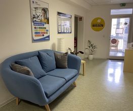 Der Eingangsbereich mit blauer Couch. An der Wand Plakate der WBS TRAINING. Rechts eine Glastür mit WBS TRAINING Beklebungen. Neben dem Sofa ein Beistelltisch mit Pflanze.