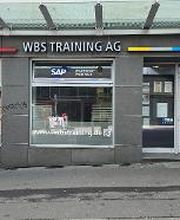 Haupteingang mit zwei großen Fenstern neben der Tür. Auf der grauen Fassade ist der Schriftzug WBS TRAINING AG angebracht.