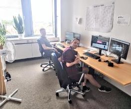 zwei Teilnehmer sitzen am Arbeitsplatz mit zwei Bildschirmen , Tastatur, Headset und Maus und arbeiten am Computer