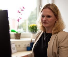Eine blonde Frau in beigem Blazer sitzt an einem Schreibtisch und schaut auf einen Monitor. Auf der Fensterbank hinter ihr stehen eine Orchidee und gelbe Blumen. Durch das Fenster ist unscharf eine grüne Umgebung zu sehen. Der Raum ist hell und ruhig.