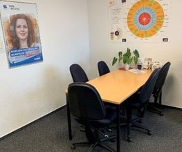 Büro bestehend aus einem langen Tisch mit mehreren schwarzen Bürostühlen. An der Wand hängen Plakate und auf dem Tisch steht eine kleine Topfpflanze.