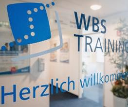 Eingangstür aus Glas mit Blick in den Flur. Auf der Tür ein blauer Schriftzug WBS TRAINING – Herzlich willkommen plus Logo.