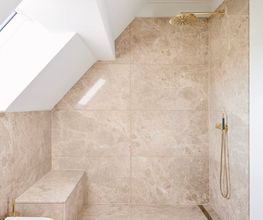 Hochwertig ausgestattetes Duschbad unter der Dachschräge mit Natursteinverkleidung, goldenen Armaturen und integrierter Sitzbank. Eine Kombination aus Funktionalität und eleganter Zurückhaltung.