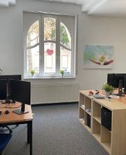 Schulungsraum mit mehreren Arbeitsplätzen mit jeweils 2 Bildschirmen, einem Bürostuhl, Tastatur, Maus und Headset. An den Wänden hängen Plakate und Bilder.