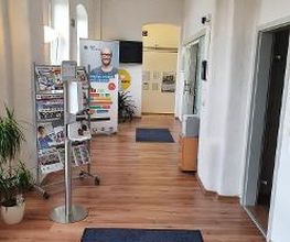 Flur zu verschiedenen Räumen. Links ein Flyerständer und dahinter ein Roll Up Banner. Rechts mehrere Türen und ein Schrank mit Drucker.