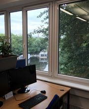 Ein Bild von einem Arbeitsbereich neben einem Fenster mit Blick ins Grüne. Der Arbeitsbereich hat blaue Trennwände auf denen ein Blumenkasten angebracht ist sowie 2 Bildschirme, Tastatur und Maus.