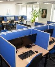 Ein Schulungsraum mit mehreren Arbeitsbereichen, die durch blaue Trennwände voneinander getrennt sind. Jeder Arbeitsplatz besteht aus 2 PC-Bildschirmen, Tastatur und Maus sowie Headset. Blaue Trennwände.