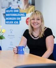 Eine Frau mit blonden Haaren und schwarzer Bluse sitzt an einem Tisch. Sie hält eine blaue Tasse in der Hand.