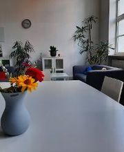 Aufenthaltsraum mit Sesseln, Sofa, Zimmerpflanzen sowie einem Tisch auf dem eine Vase mit bunten Blumen steht.