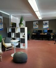 Schulungsraum mit mehreren Arbeutsplätzen mit jeweils 2 Bildschirmen, einem Bürostuhl, Tastatur und Maus. Links stehen 2 Sessel und ein Beistelltisch. An der Wand hängen 3 Plakate. Ein Regal trennt den Sitzbereich von der offenen Tür.