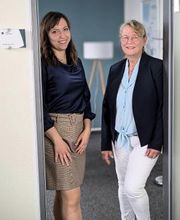 2 Mitarbeiterinnen stehen in einer Tür und lachen freundlich in die Kamera.