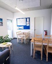 Aufenthaltsraum mit Tischen, Stühlen, einem Sofa und zwei Fenstern. An den Wänden hängen eine blaue Pinnwand mit Informationen und mehrere Leinwand Bilder. 