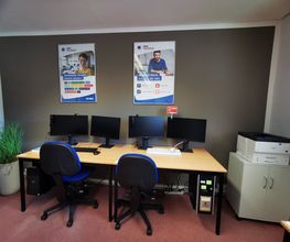 Arbeitsplätze mit jeweils einem Bürostuhl, 2 Bildschirmen und Tastatur und Maus. Links eine Zimmerpflanze, rechts ein Regal mit Drucker. An der Wand 2 Plakate der WBS TRAINING.