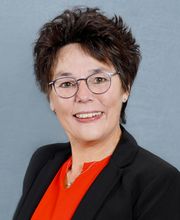 Eine Frau mit kurzen dunklen Haaren und Brille. Sie trägt einen schwarzen Blazer und eine rote Bluse.