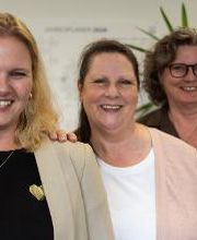 Drei lächelnde Frauen stehen nebeneinander. Links trägt eine Frau Beige mit Herz-Anstecker, in der Mitte Weiß mit rosa Jacke, rechts Dunkel mit Brille. Im Hintergrund sind eine helle Wand, Pflanzen und verschwommen ein Jahresplaner zu sehen.