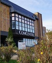 Außenansicht vom Eingangsbereich des Gebäudes. Das Gebäude hat eine Fensterfront und zeigt den Schriftzug „COMCENTER“.
