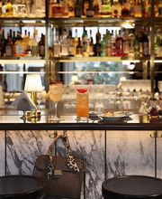 Marble bar counter set with elegant cocktails, a small table lamp, and delicate bites, backed by shelves of illuminated spirits for a refined aperitivo atmosphere.
