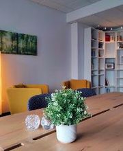 Gemütlicher Aufenthaltsraum mit 2 gelben Sesseln, Stehlampe, Bücherregal, großem Holztisch mit blauen Stühlen und Topfpflanze sowie Dekoration. Ein Bild von einem Wald hängt an der Wand.