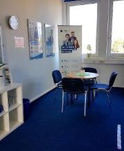 Pausenbereich mit links einem Regal mit Zimmerpflanze, mittig ein Tisch mit 4 Stuhlen. Dahinter ein Banner und Fenster mit Pflanzen. An der Wand hängen 2 Plakate.