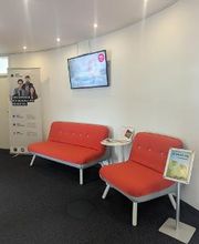 Wartebereich im Flur mit 2 modernen, roten Sesseln, einem dazugehörigen Beistelltisch, einem Fernseher an der Wand und einem Werbeaufsteller im Hintergrund.