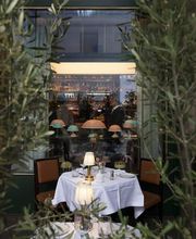 Intimate outdoor table set with white linens and a small table lamp, framed by potted olive trees and reflected against a softly lit bar interior behind the window.