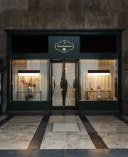 Exterior storefront of Sant Ambroeus with dark green façade, gold-trimmed signage above the entrance, softly lit display windows showcasing pastries and floral arrangements, and sheer curtains framing the doorway against a stone-lined arcade.