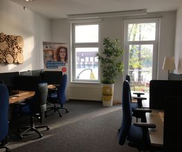 Schulungsraum mit mehreren Arbeitsbereichen, die durch blaue Trennwände voneinander abgegrenzt sind und mit 2 PC-Bildschirmen, Tastatur, Maus und Headset ausgestattet sind. Zugang zum Balkon.