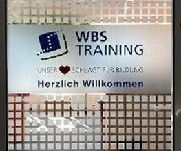 Glastür im Eingangsbereich. Schild mit Aufschrift „WBS TRAINING – Unser Herz schlägt für Bildung – Herzlich Willkommen“