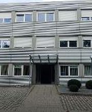 Ein dreistöckiges Bürogebäude mit grauer Metallfassade und vielen Fenstern. Der Eingang in der Mitte hat eine Glasüberdachung. Vor dem Gebäude ist ein gepflasterter Parkplatz mit einem schwarzen Auto und einem Hinweisschild. Büsche schmücken den Eingang.