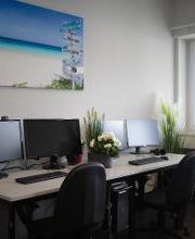 Zwei Arbeitsplätze für Teilnehmedne mit 2 Bildschrimen, Tastatur, Maus und Headset. An der Wand hängt ein schönes Strandbild und auf den Tischen stehen Blumen.