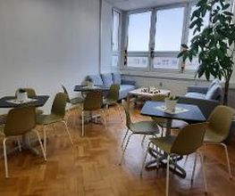 Pausenraum mit Fensterfront und 3 Tischen mit jeweils 4 grünen Stühlen. Im Hintergrund stehen sich 2 Sofas gegenüber, zwischen ihnen ein Beistelltisch. Rechts steht eine große Zimmerpflanze.