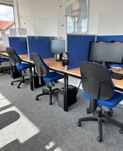 Schulungsraum mit 3 Arbeitsplätzen, jeweils mit einem Bildschirm, einem Bürostuhl, Tastatur, Maus und Headset. Blaue Trennwände grenzen die Tische voneinander ab.