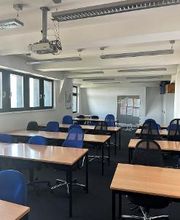 Schulungsraum mit Tischreihen an denen blaue Bürostühle stehen. An der Wand hängt ein Beamer und im Hintergrund gibt es eine Moderationstafel.