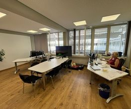 Büroraum mit 3 Schreibtischen vor einer großen Fensterfront. An einem Schreibtisch sitzt eine Mitarbeiterin vor ihrem PC-Bildschirm.