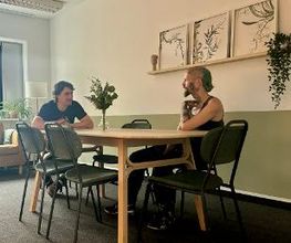 Pausenraum mit langem Tisch und 6 Stühlen. Im Hintergrund ein gelbes Sofa und an der Wand ein Regal mit 3 Bildern und einer Zimmerpflanze. 2 Personen sitzen am Tisch und unterhalten sich.