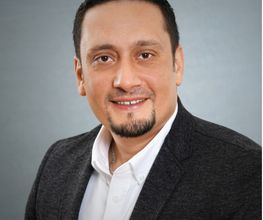 Mann mit kurzen, schwarzen Haaren. Er trägt ein Graues Sakko und ein weißes Hemd.
