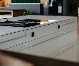Die Detailaufnahme zeigt eine hochwertige Kücheninsel mit eleganten weißen Auszügen, schlanken Designgriffen und einer modernen Arbeitsplatte in Betonoptik. Im Mittelpunkt steht ein flächenbündig integriertes Induktionskochfeld mit Muldenlüfter, ergänzt durch eine Grillplatte für vielseitige Kochmöglichkeiten. Die Kombination aus klaren Linien, hochwertigen Materialien und durchdachter Funktionalität macht diese LEICHT Designküche zu einem Highlight moderner Küchenplanung im Küchentreff Liebold.