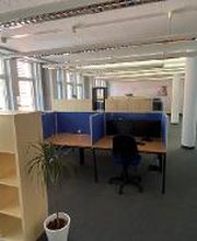 Schulungsraum mit mehreren Arbeitsplätzen und einem Holzregal mit Pflanzen. Der Raum hat mehrere Fenster und Säulen im Gang.