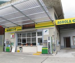 AGROLA Tankstelle in Gersau unter Wellblechdach; zwei Tanksäulen; Spitex Logo an Fassade über Tanksäule; Fensterfront und Tür zwischen Säulen it Blumentöpfen auf Fensterfront