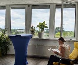 Eine Frau sitzt in einem gelben Schwingsessel und liest etwas. Vor ihr steht ein Bistrotisch mit blauer Husse.