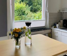 Küche mit Tisch und Stühlen. Auf dem Tisch steht eine kleine Blumenvase und eine Wasserflasche.