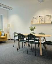 Pausenraum mit langem Tisch und 6 Stühlen. Links ein gelbes Sofa und eine Stehlampe. An der Wand ein Regal mit 3 Bildern und einer Zimmerpflanze.