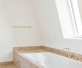 Elegantes Bad mit Einbaubadewanne in Natursteinfassung. Die Kombination aus klarer Formensprache, hellen Materialien und goldenen Armaturen schafft eine luxuriöse, zeitlose Atmosphäre.