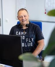 Ein Mitarbeiter sitzt in einem Büroraum an seinem Schreibtisch vor dem PC-Bildschirm. Er trägt ein Headset um den Hals und schaut freundlich in die Kamera.