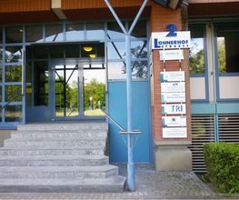 Außenansicht des Eingangsbereich vom Gebäude. Glas Eingangstür mit blauen Rahmen. Rechts an der Seite hängt eine Schildertafel der ansäßigen Unternehmen.