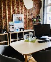 Modern und gemütlich gestalteter Beratungsraum. Weißer Tisch, graue Lounge-Sessel. Vor der Backsteinwand flache Regale mit Infomaterial. Großes Sprossenfenster. Parkettfußboden.