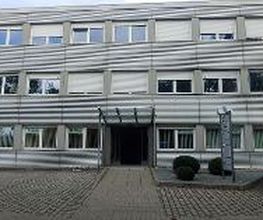 Ein dreistöckiges Bürogebäude mit grauer Metallfassade und vielen Fenstern. Der Eingang in der Mitte hat eine Glasüberdachung. Vor dem Gebäude ist ein gepflasterter Parkplatz mit einem schwarzen Auto und einem Hinweisschild. Büsche schmücken den Eingang.