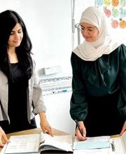 2 Frauen stehen im Schulungsraum an einem Schreibtisch und schauen sich Unterlagen in einem Ordner an. Eine Frau trägt ein Brille und ein weißes Hijab.